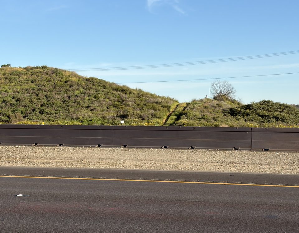 Desert hillside with wildlife crossing fencing and Natina-treated natural-looking roadside Zoneguard barrier along a highway corridor.